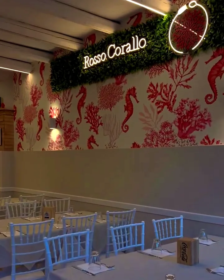 La sala di Rosso Corallo: tavoli apparecchiati, sedie bianche, coralli e cavallucci marini sulla parete e l'insegna luminosa col nome